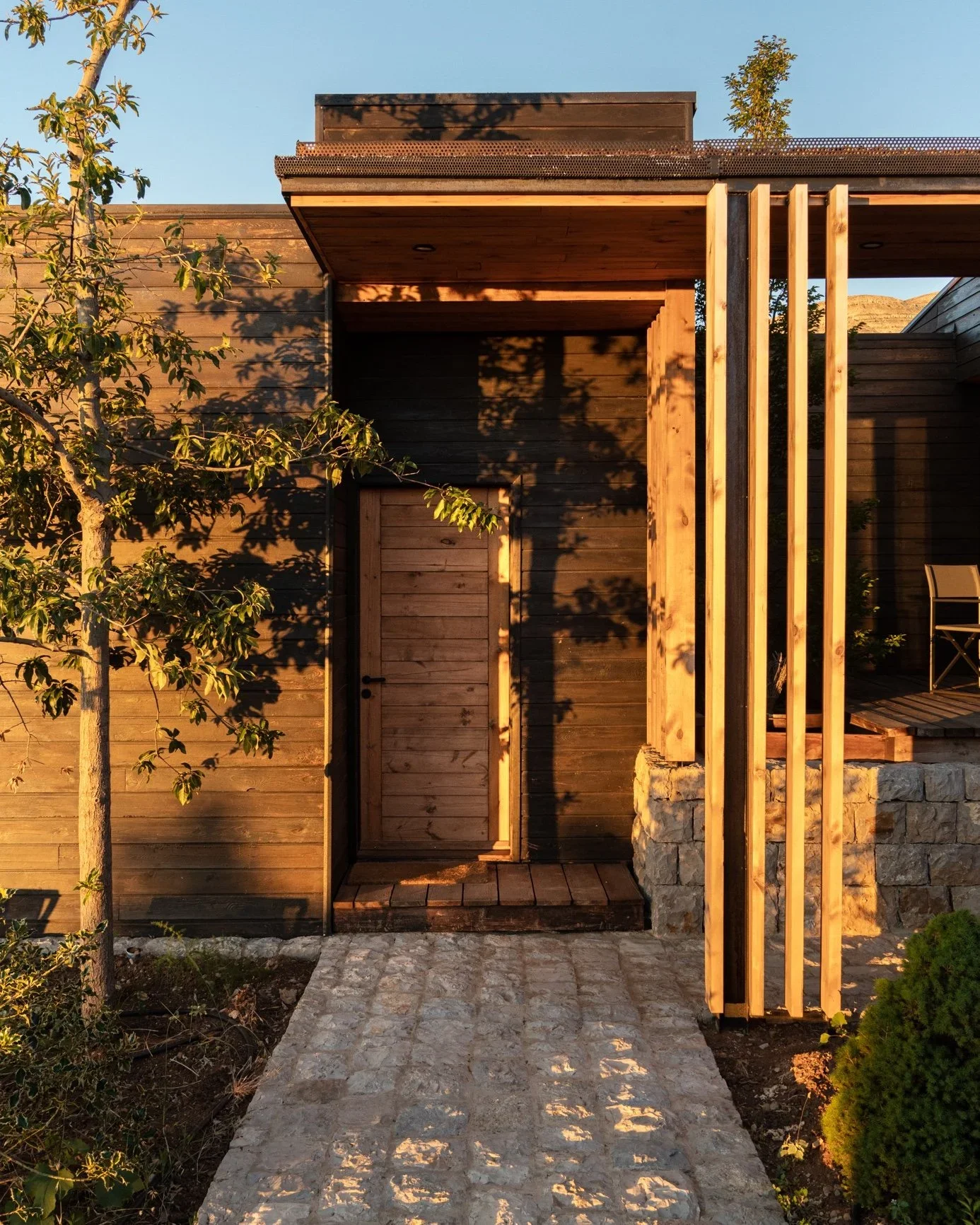 Modern wooden house exterior with a small pathway, greenery, and sunlight casting shadows.