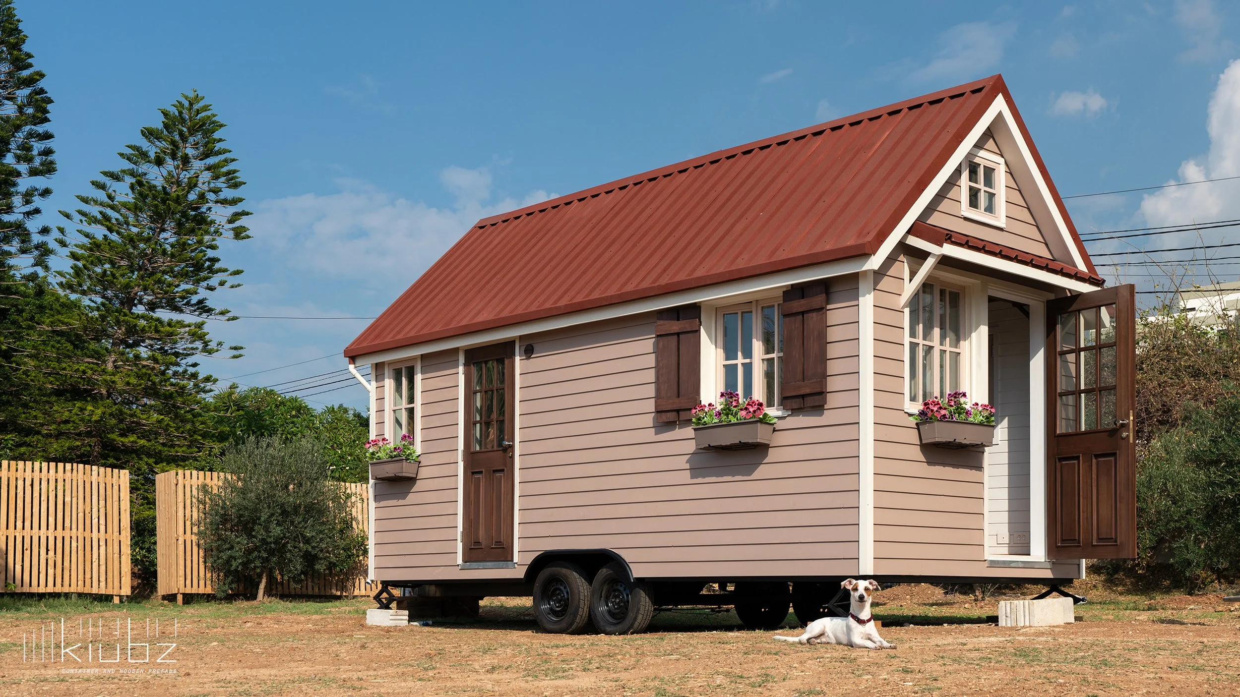 A tiny house on wheels with beige siding, a red metal roof, and wooden window shutters, situated outdoors with trees and a clear blue sky in the background, and a white dog lying on the grass.