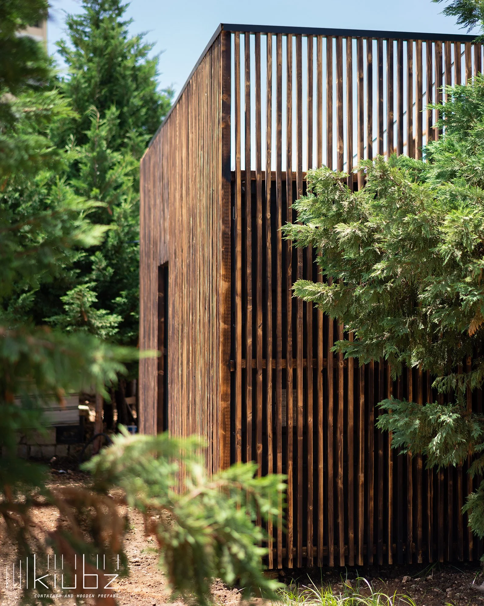 A modern wooden structure with vertical slats, partially obscured by green foliage, in an outdoor setting with clear blue sky.