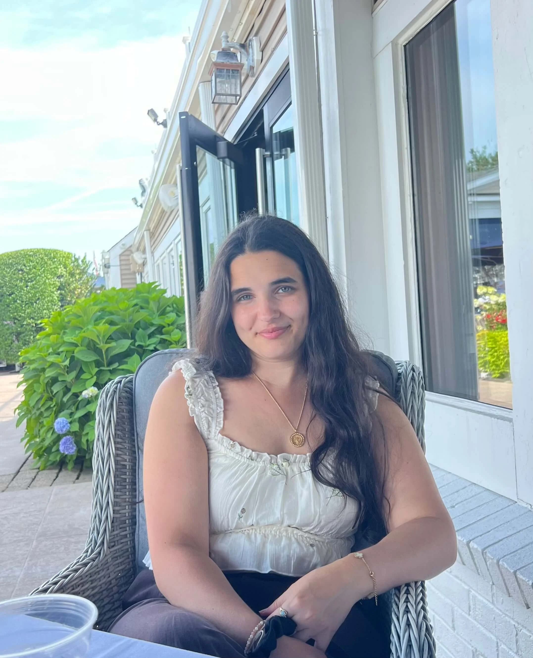 Founder Julianne Baron wearing a sleeveless white blouse and gold coin necklace, sitting outside at a restaurant.
