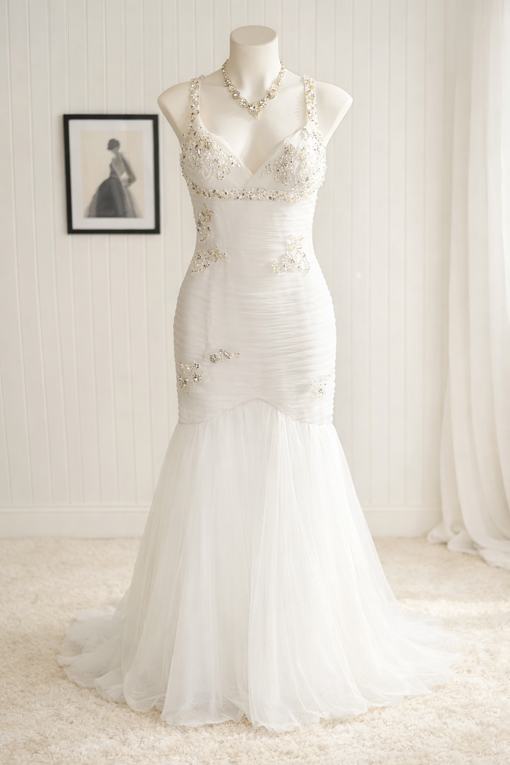 A white wedding dress with floral embellishments, displayed on a mannequin in a softly lit room, with a framed black-and-white photo of a woman in a gown hanging on the wall behind it.