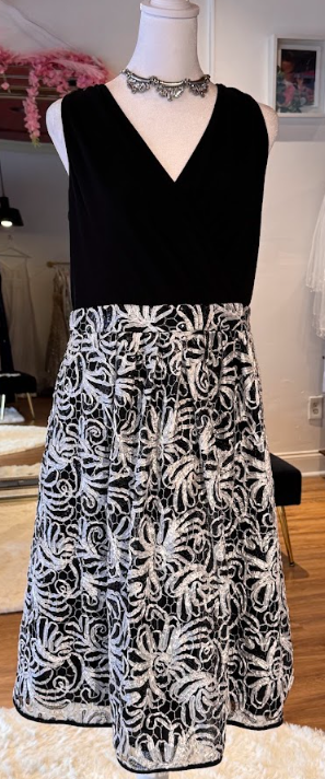 Black sleeveless dress with a V-neckline and a white lace skirt featuring floral and leaf patterns. The dress is displayed on a mannequin with a silver necklace.