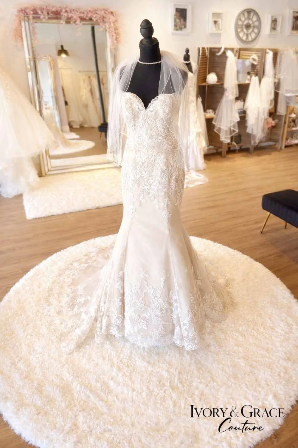 A wedding dress on a mannequin in a bridal boutique, displayed on a round fluffy rug. The dress is white with lace details, and the boutique has a mirror, a large clock, and shelves with bridal accessories in the background.