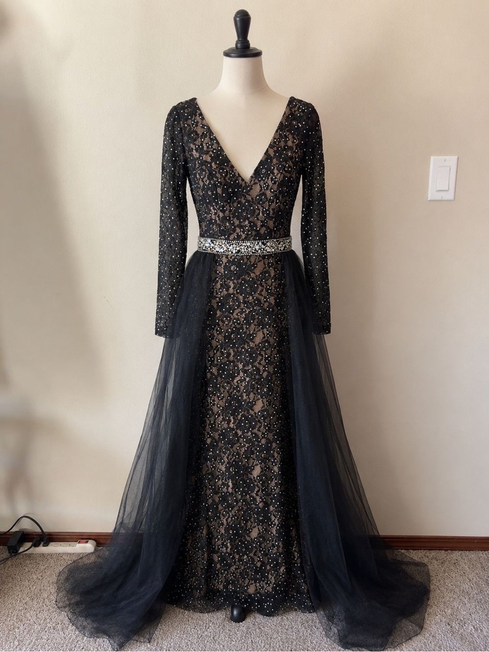 A long black evening gown with lace and sequin embellishments, featuring a deep V neckline, long sleeves, a fitted waist with a sparkly belt, and a sheer tulle overlay at the bottom.