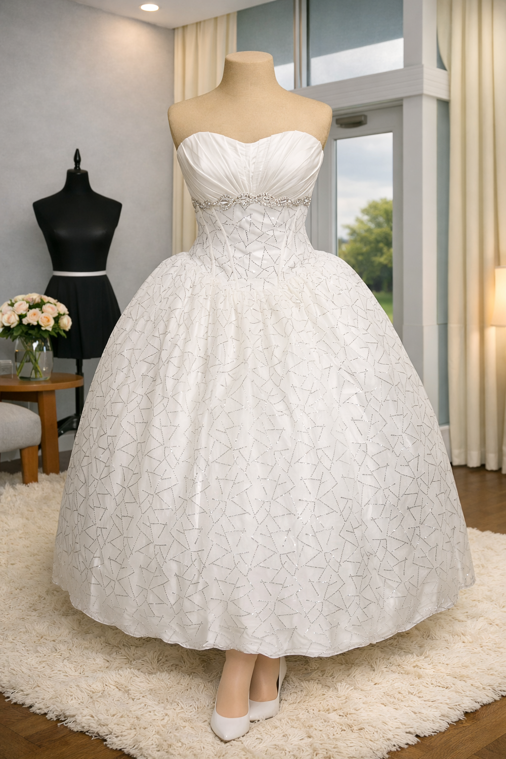 A white wedding dress on a mannequin in a well-lit room with beige curtains, a window, and a cream-colored rug. The dress features a strapless sweetheart neckline with ruched bodice, an embellished waistline, and a full skirt with a star pattern.