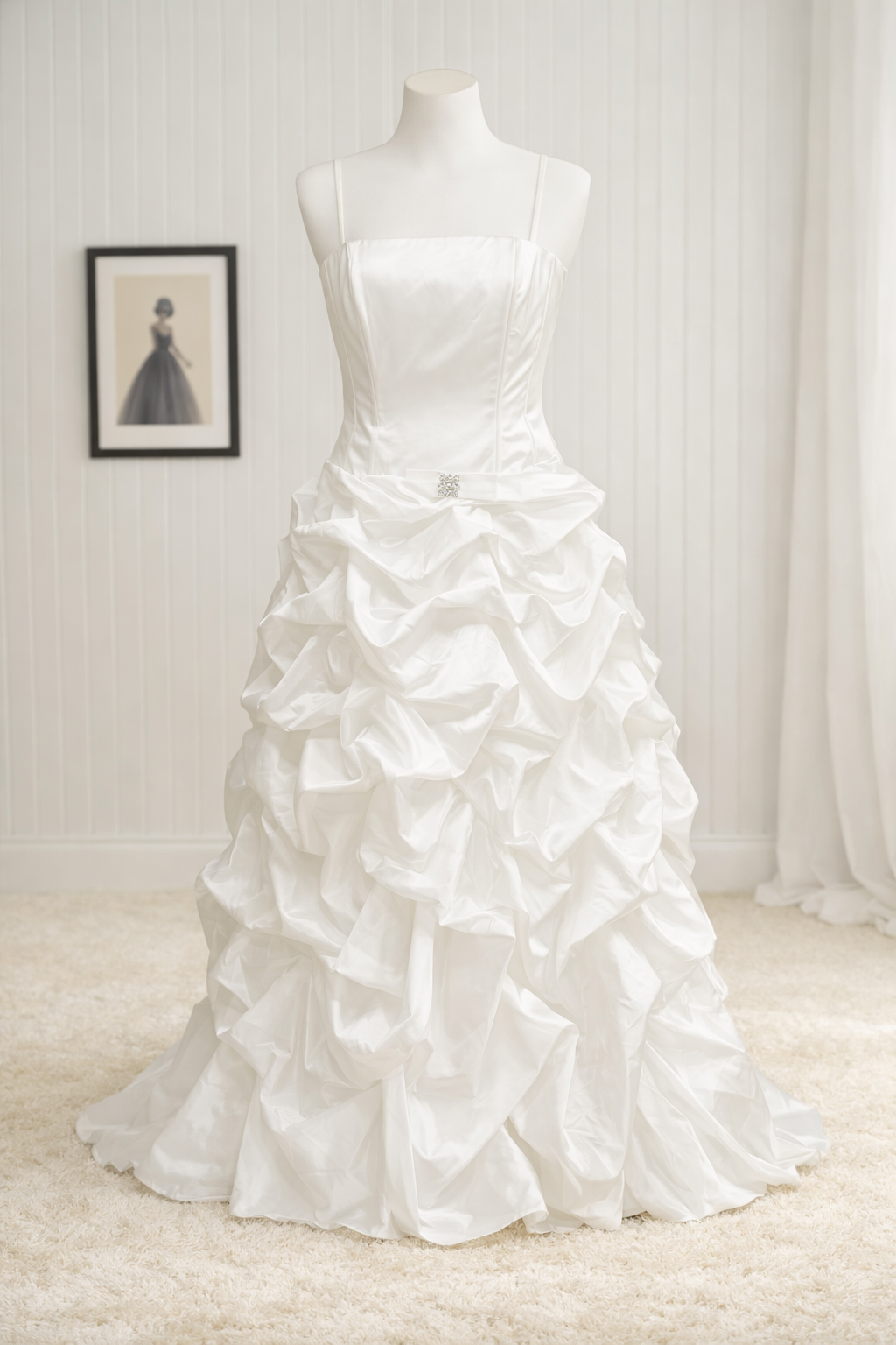 A white wedding gown with a fitted bodice and prominent ruffled skirt displayed on a mannequin in a room with white paneled walls and a framed fashion illustration in the background.