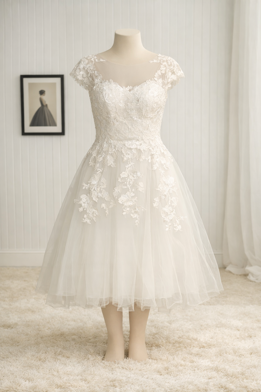 A wedding dress on a mannequin, featuring lace and tulle fabric with floral embroidery details, in a room with light-colored walls and a framed photo in the background.