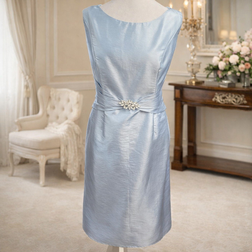 A light blue satin dress on a mannequin in a luxurious room with cream-colored walls, a white armchair with a blanket, a wooden sideboard with floral decorations, and a mirror.