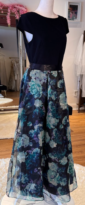 A mannequin wearing a black top with short sleeves and a long, floral, semi-transparent skirt with shades of blue, gray, and black, displayed in a boutique with clothing racks and a mirror in the background.