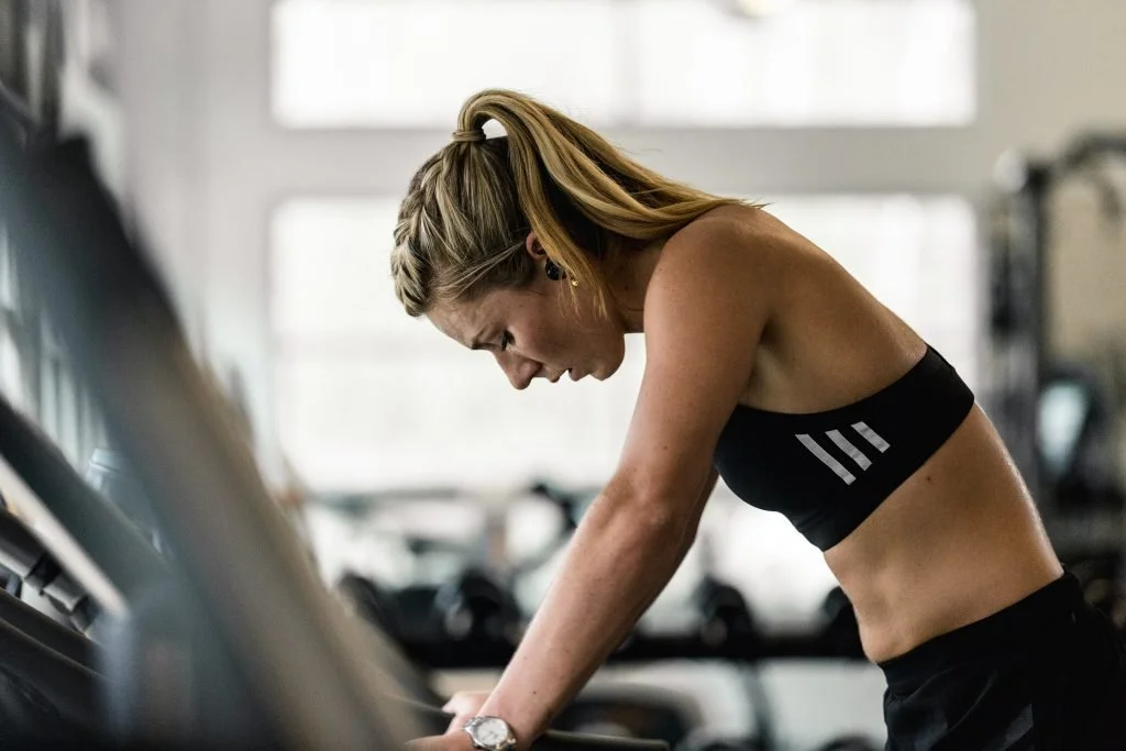 A woman skier with blonde hair in a braid, wearing a black sports bra with white stripes, is leaning forward and working on a treadmill in a gym.