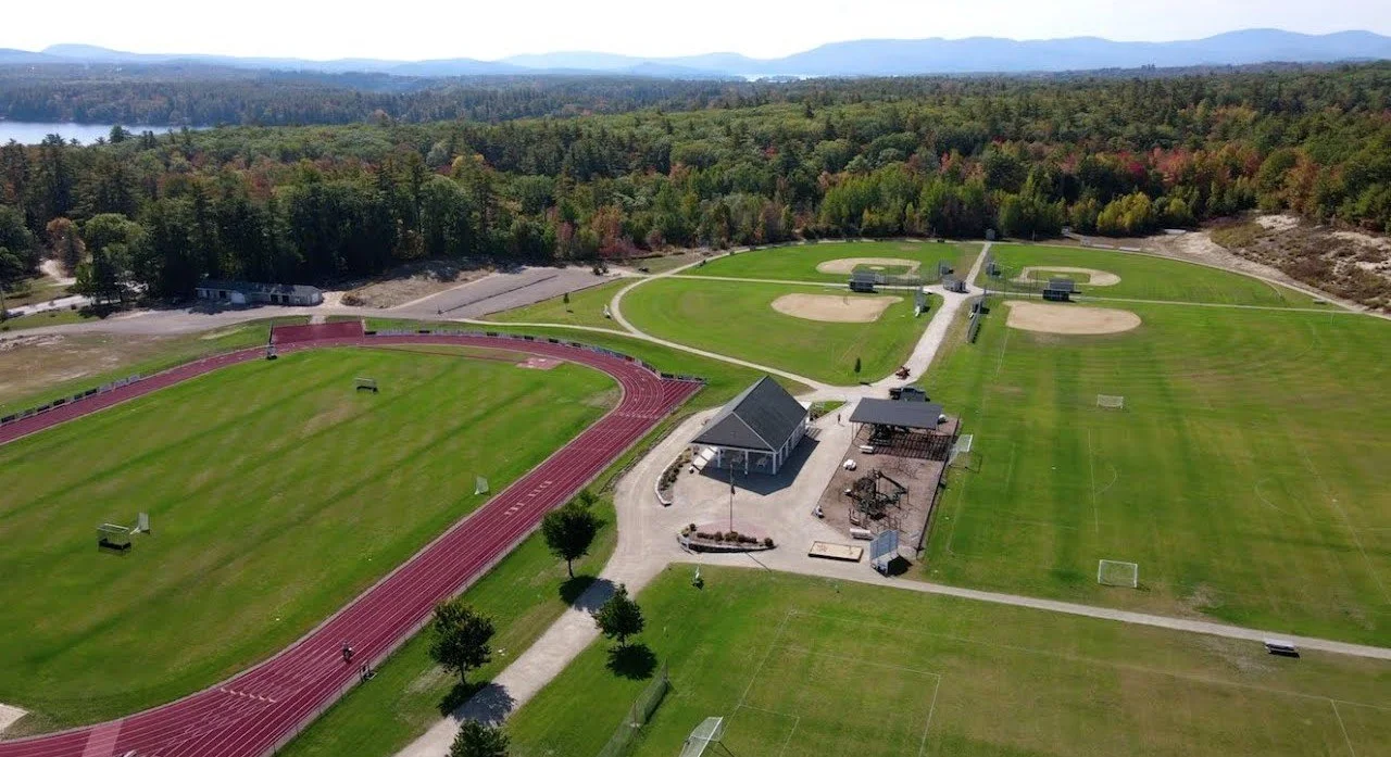Aerial view of a sports complex called The Nick located in Wolfeboro, NH with a running track, baseball diamonds, soccer fields, and a small park surrounded by trees and hills.