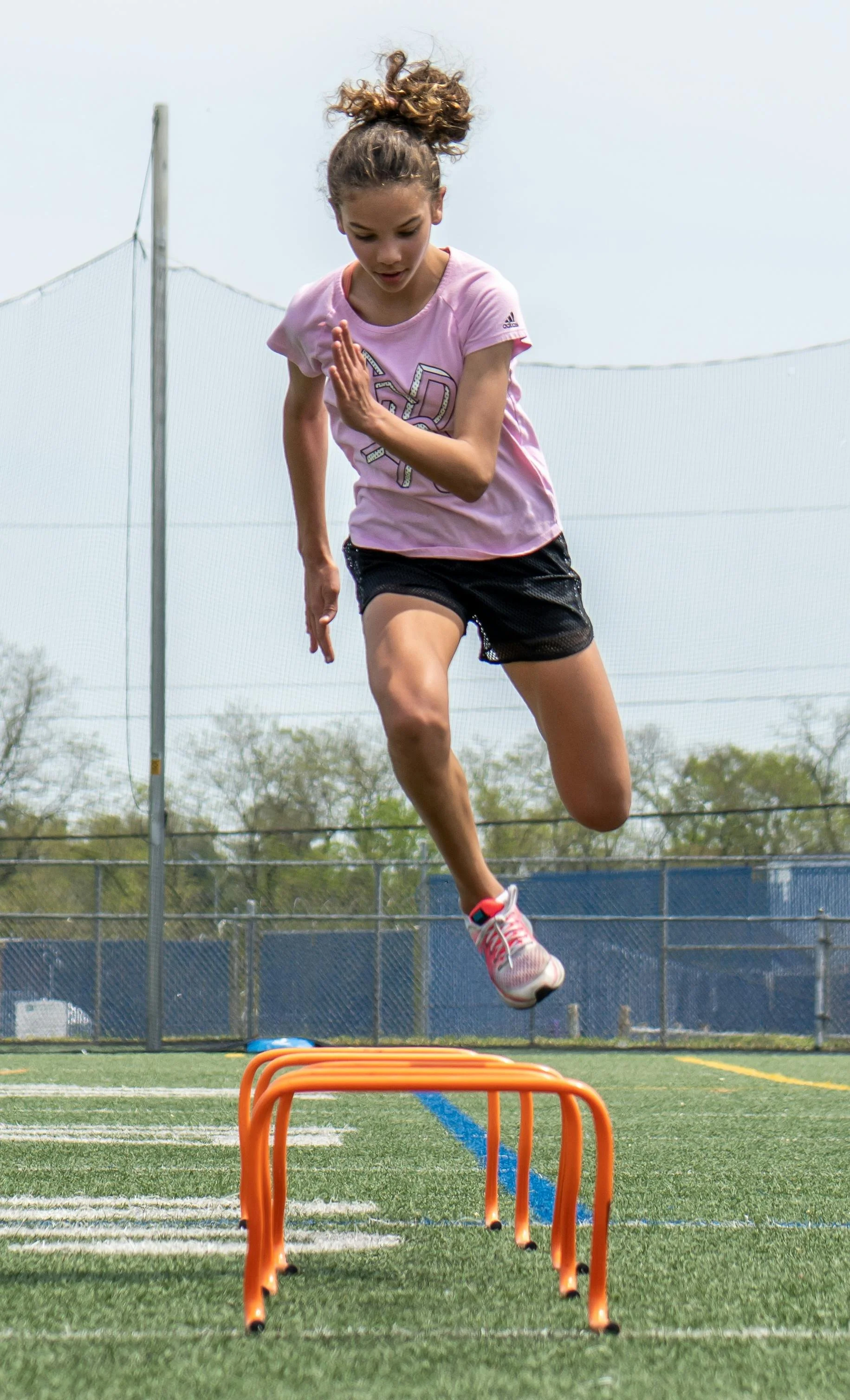 A girl in pink shirt and black shorts jumping over orange hurdles on a grassy track.