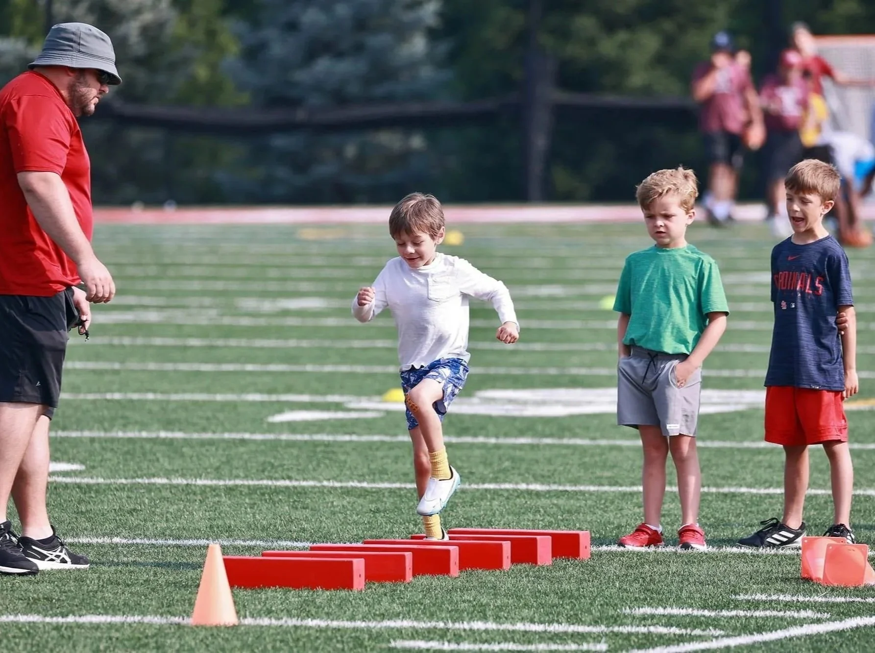 A man coaching children during an outdoor athletic event on a field, with a boy jumping over an obstacle, and other children waiting.