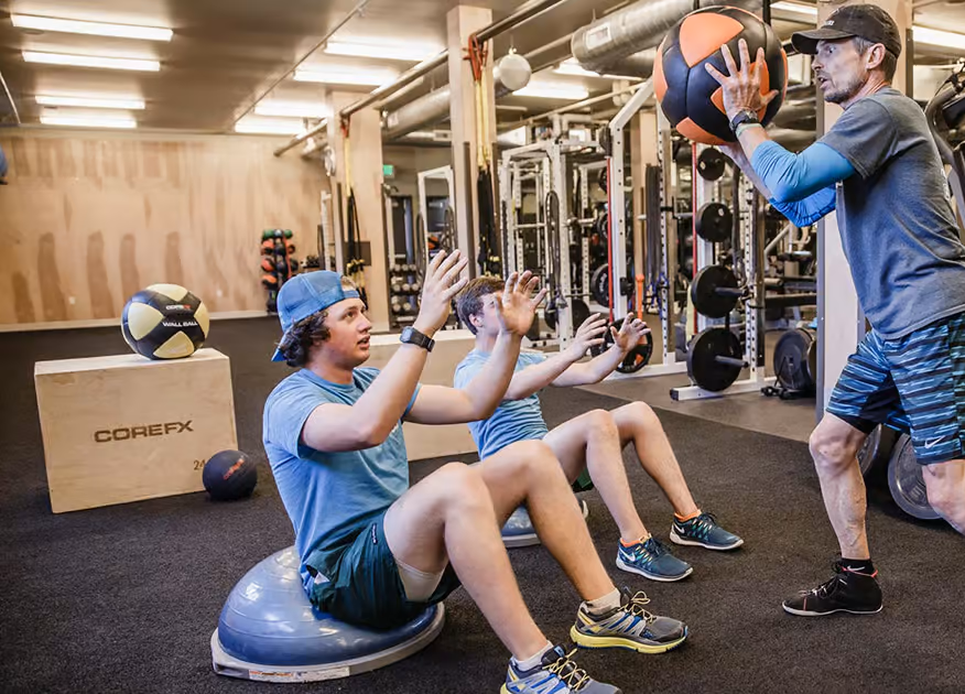 A man holding a medicine ball in a gym with two young boys sitting on exercise balls in front of him, reaching out with their hands.