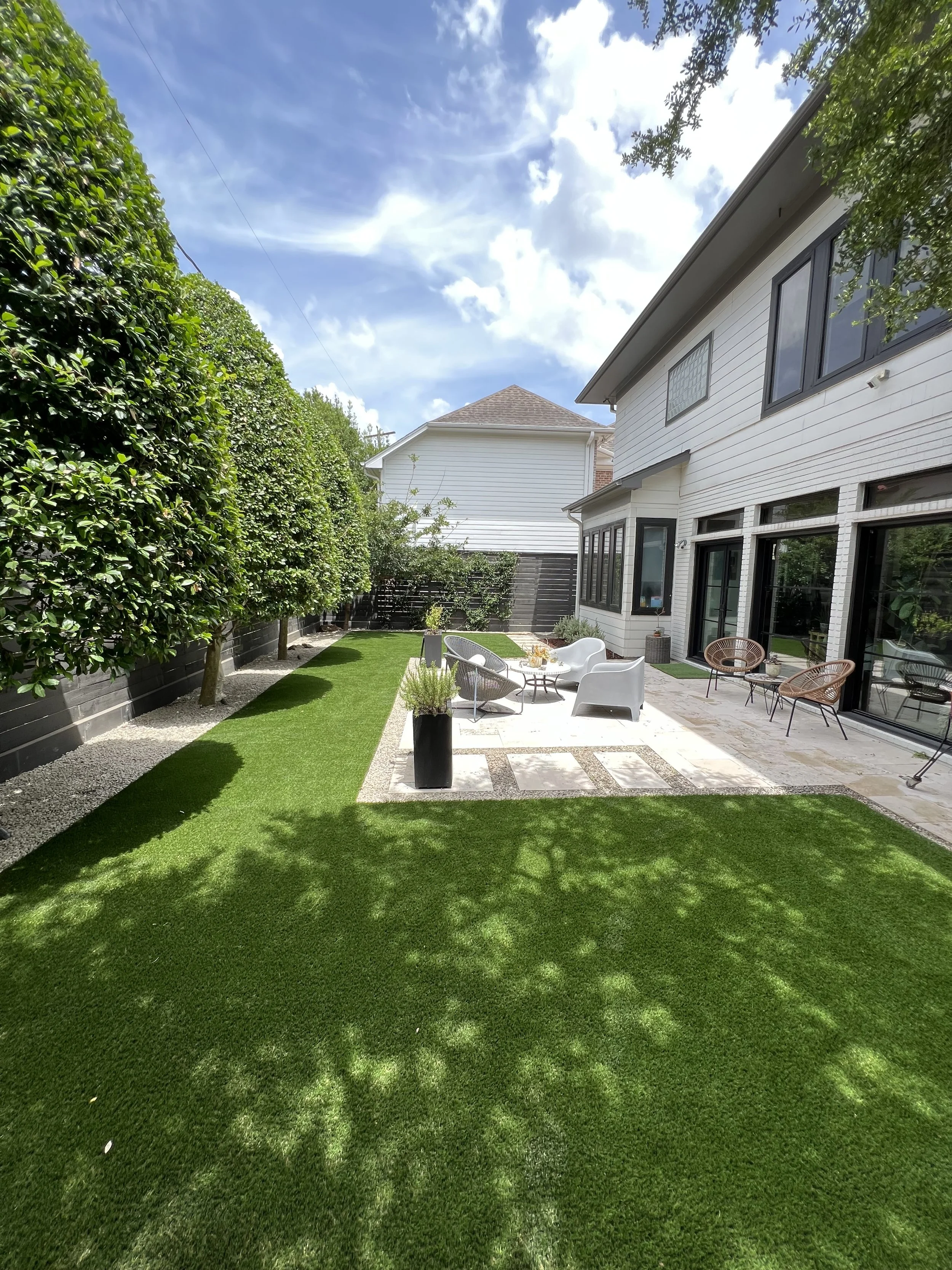 Backyard with neatly trimmed green grass, patio area with outdoor furniture, tall hedge on the left, house with large windows on the right, blue sky with clouds overhead.