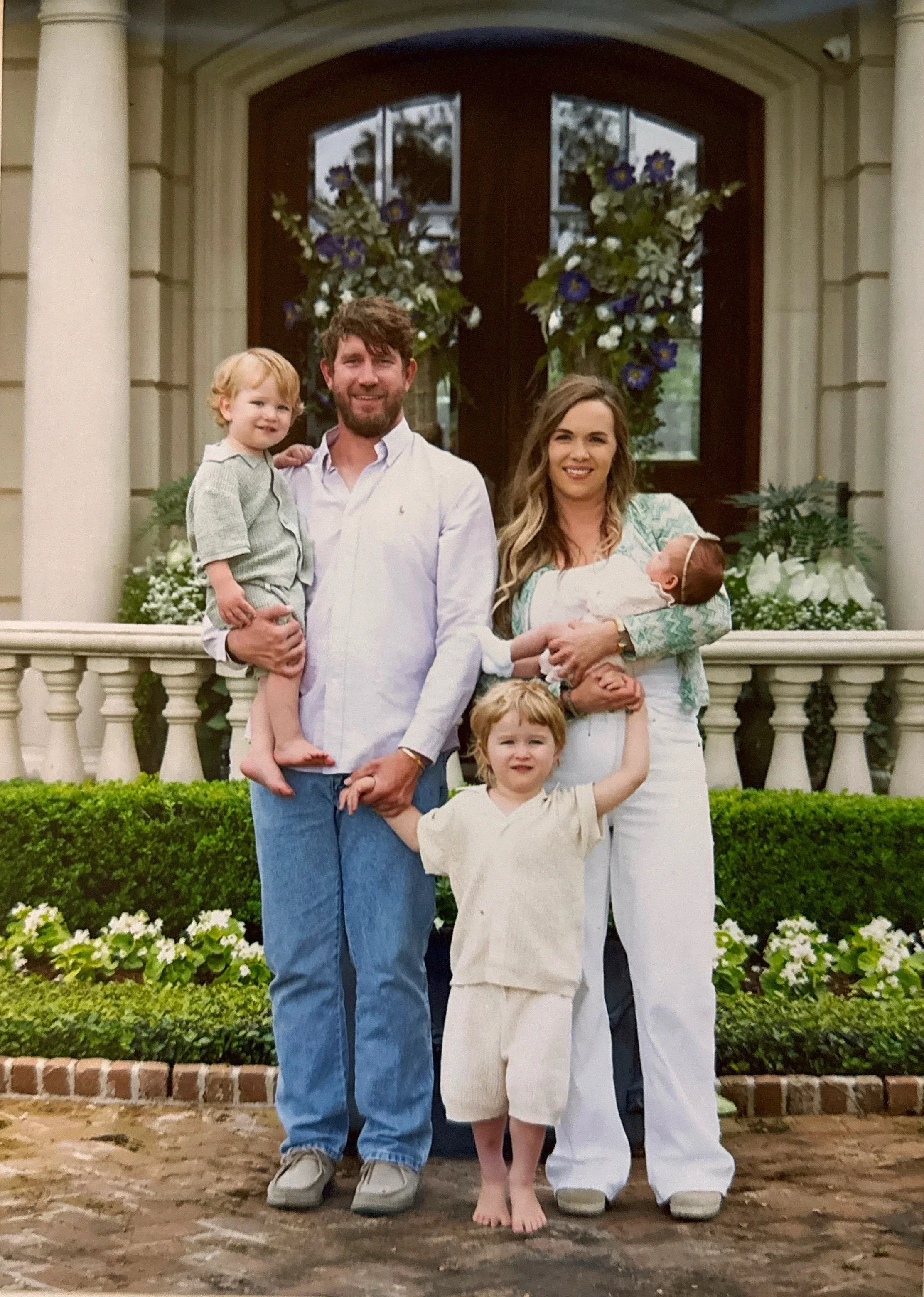 Family portrait of four people standing on a porch in front of a large wooden door with glass panels, decorated with flowers. Two children, a man, and a woman are smiling and posing for the photo.