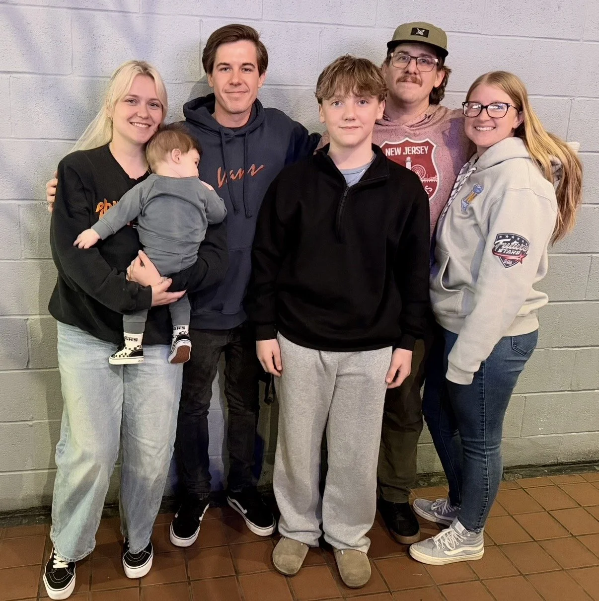 A group of six people standing against a gray brick wall, including four adults, one teenager and a small child, with the small child being held by a woman on the far left.
