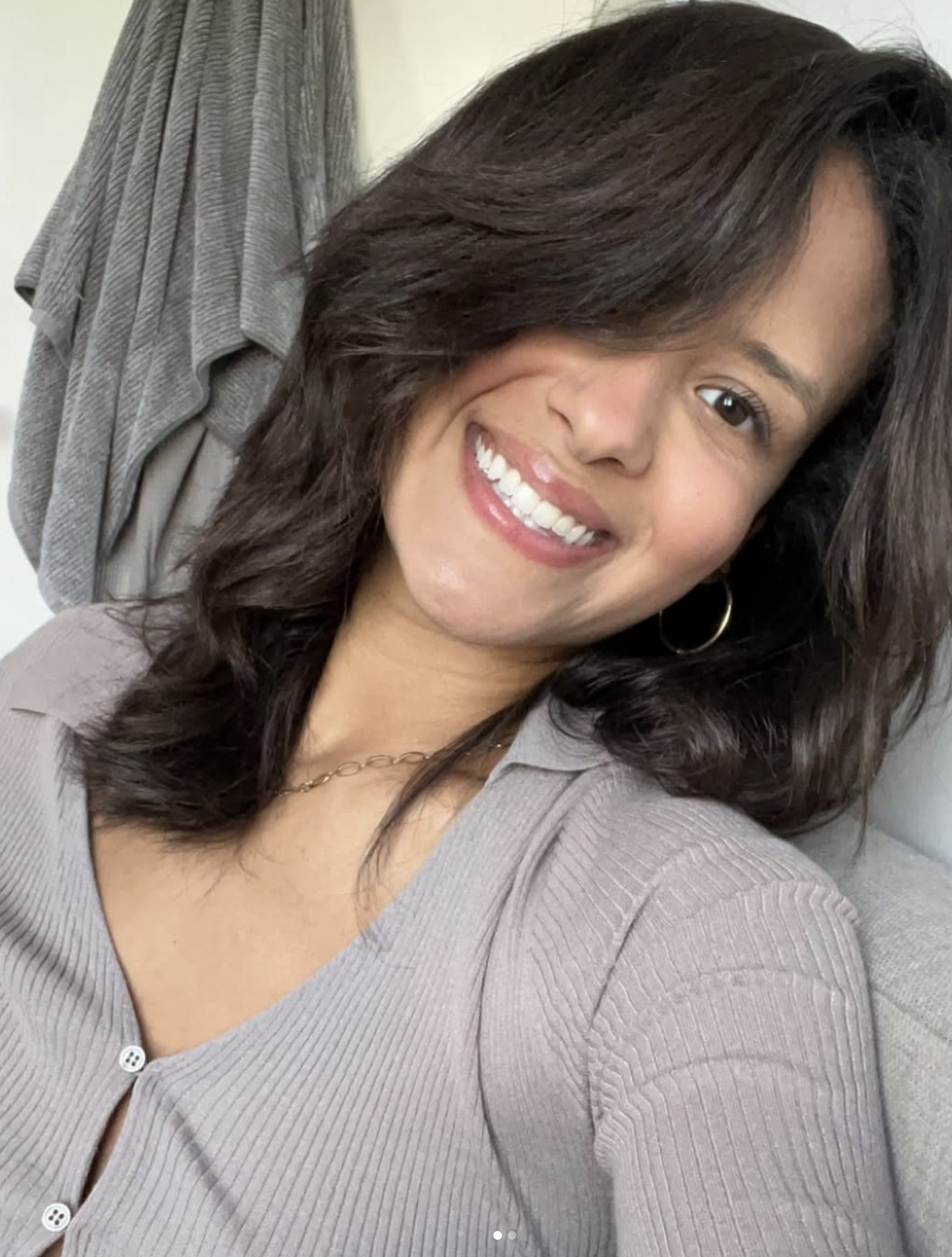 A photo of Hannah with dark, wavy hair taking a selfie indoors, smiling, wearing a beige button-up top.