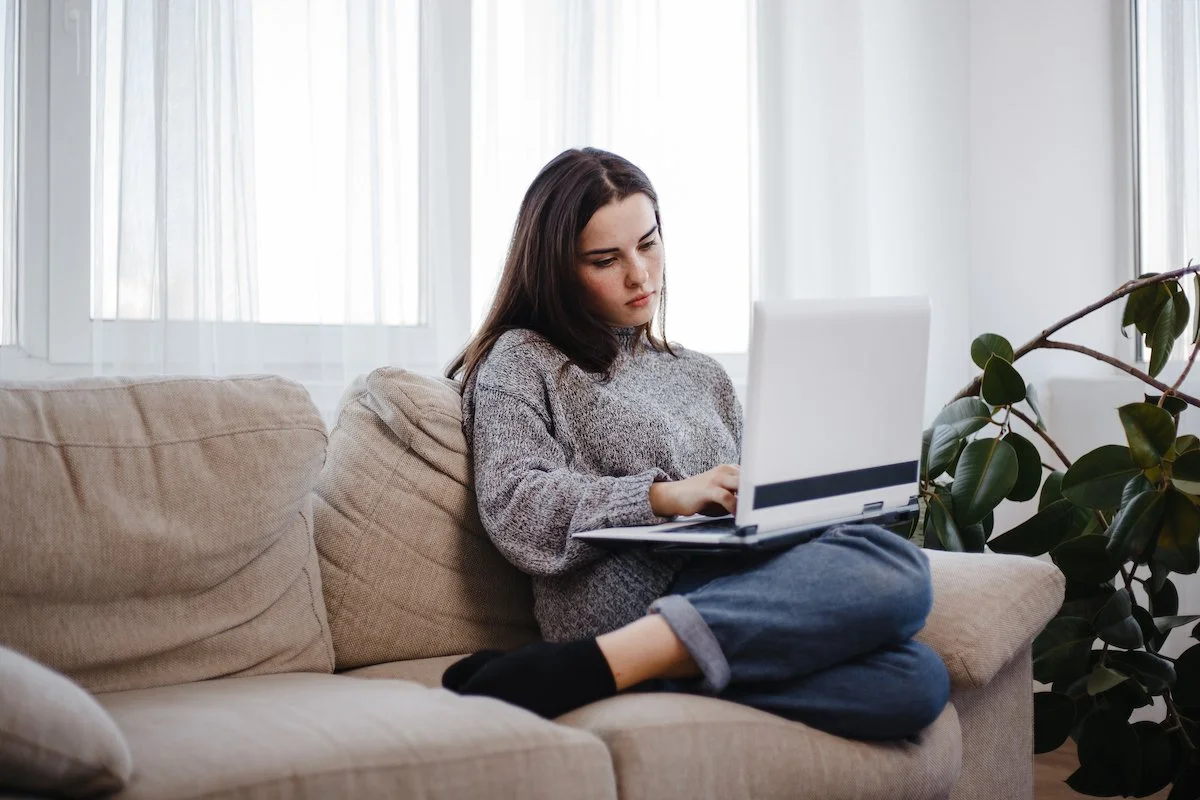 Young woman sitting on a beige couch working on a laptop, with natural light coming through windows behind her, and a potted plant nearby.