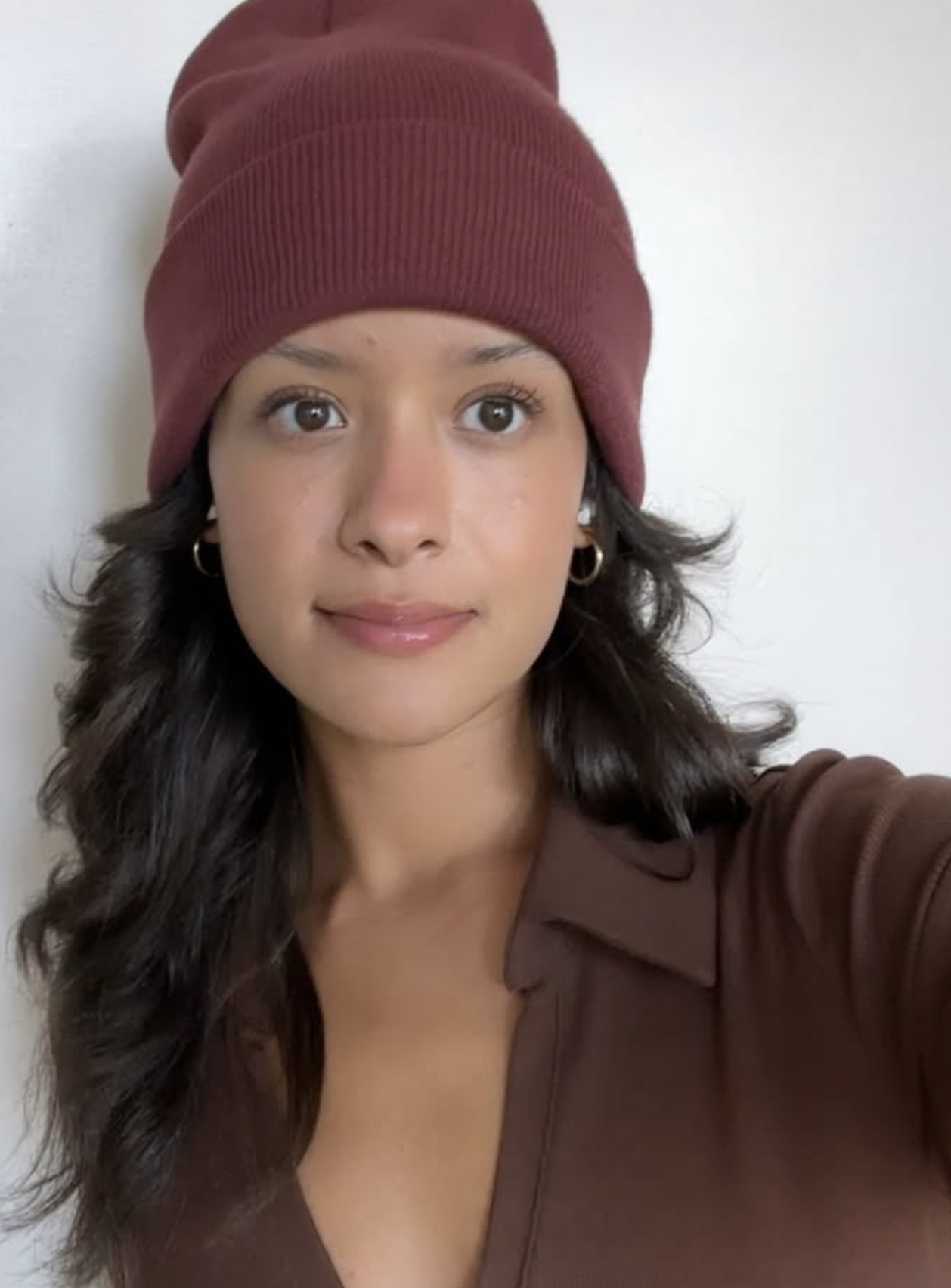 A photo of Hannah with long wavy dark hair wearing a maroon beanie and brown shirt, standing against a plain white background.