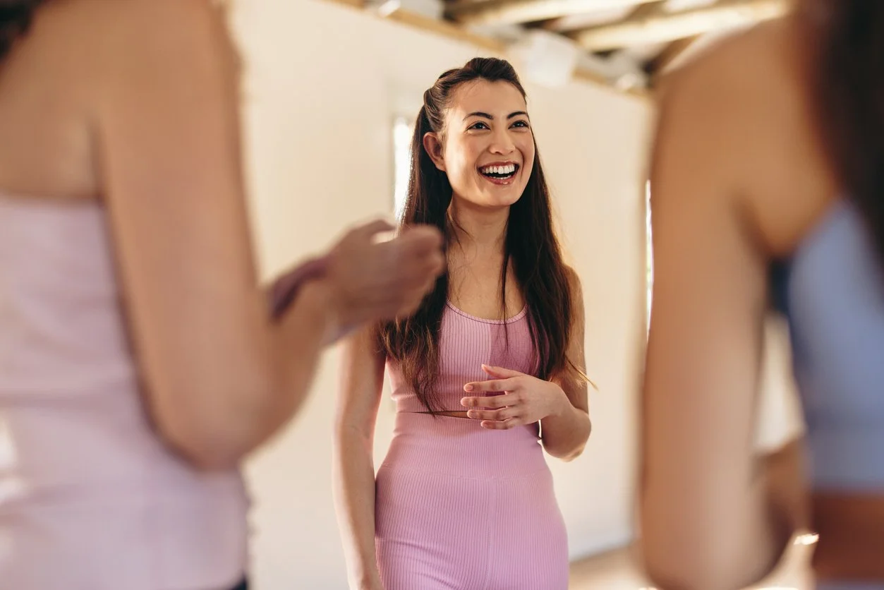 A woman in a pink sports bra and leggings, smiling and laughing surrounded by two other women in workout clothes.