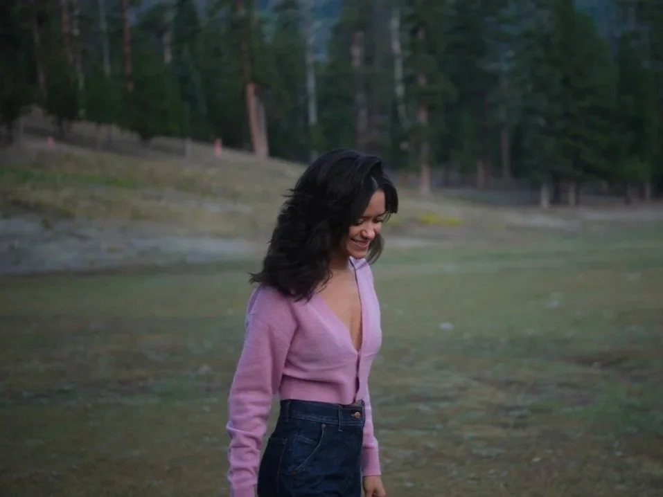 A woman with dark hair wearing a pink cardigan and dark jeans, smiling and looking down, standing in a grassy outdoor area with trees in the background.