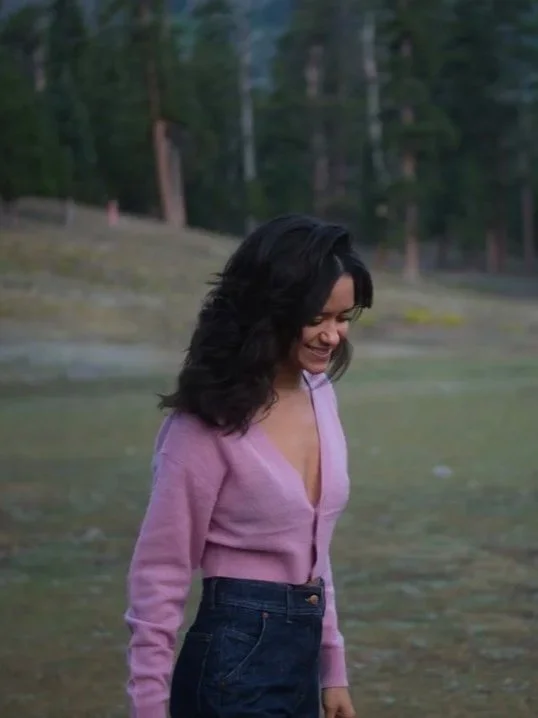 A woman with shoulder-length dark hair smiling and looking down, wearing a pink cardigan and dark jeans, outdoors with a forested background.