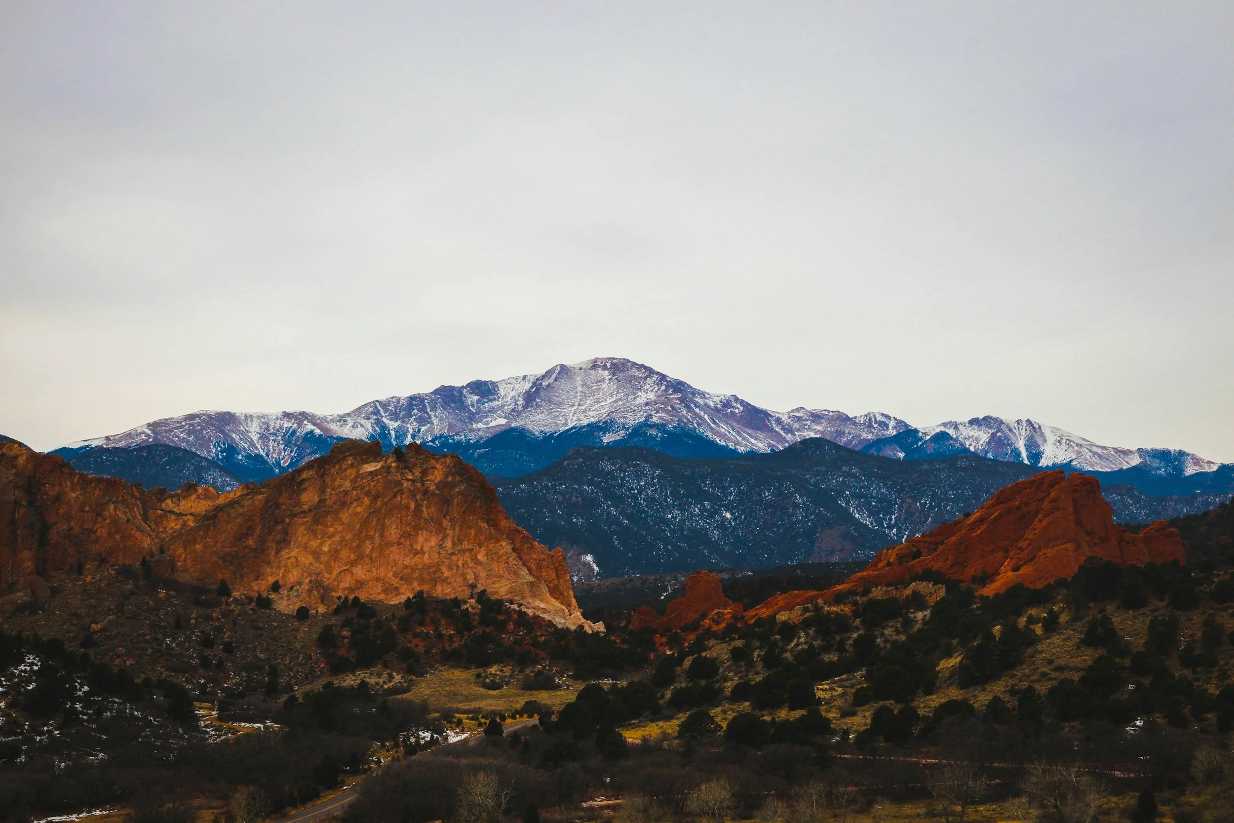 Mountain landscape with reddish rocky formations in the foreground and snow-capped mountains in the background under an overcast sky.