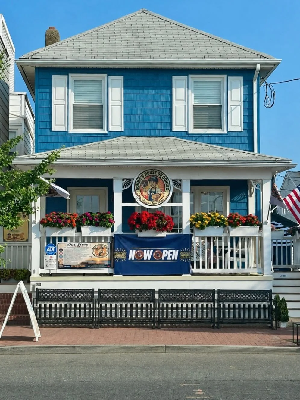 Blue two-story house with a front porch decorated with flower boxes and a sign that says "NOW OPEN," offering donuts. The house features white window shutters and a sign above the porch that reads "Sonny's House of Donuts."