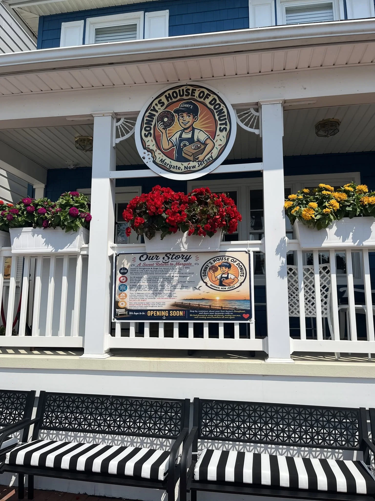 Exterior of Sonny’s House of Donuts in Margate, New Jersey with a sign featuring a cartoon boy holding a donut and a box of donuts, surrounded by flower pots with red and yellow flowers, and two black and white striped benches in front.
