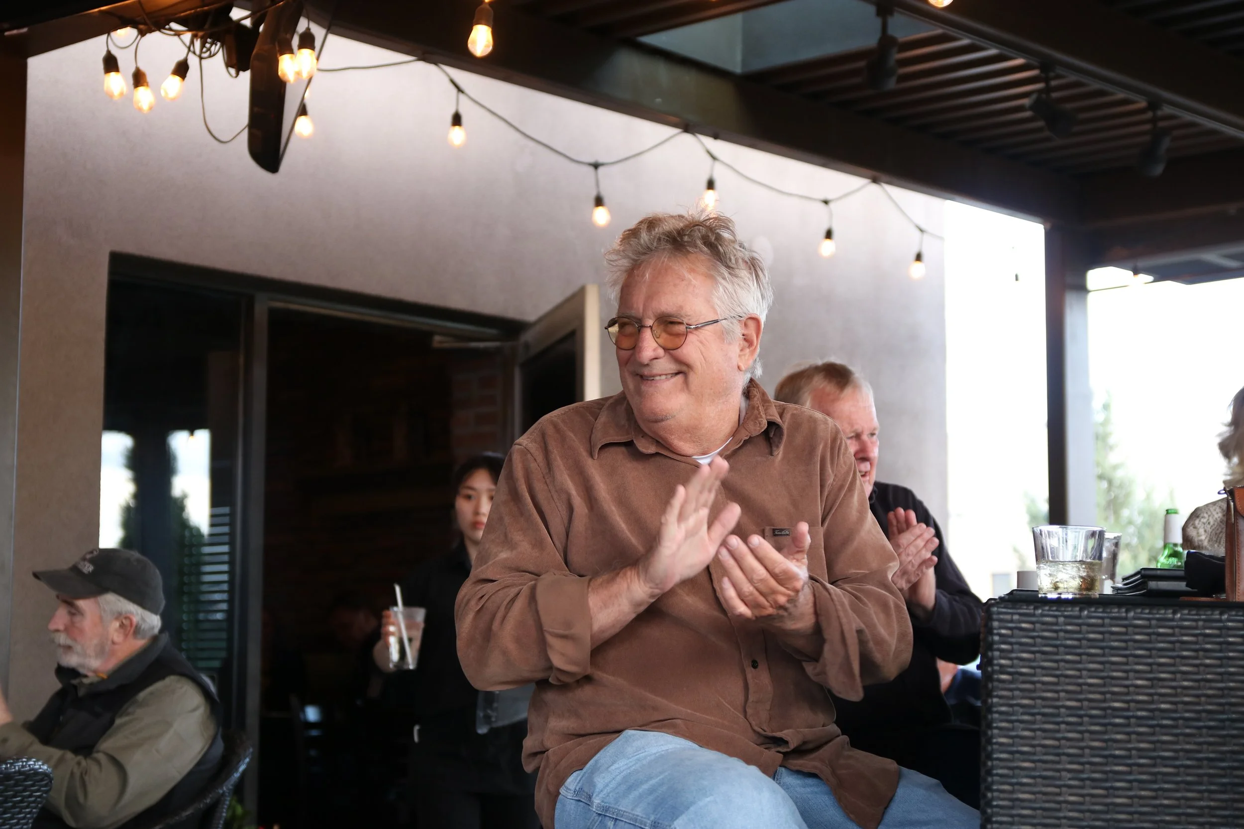 A group of people enjoying themselves at an outdoor gathering under string lights, with a smiling man in the center clapping.