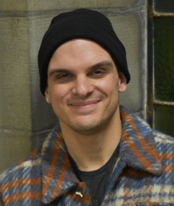 A smiling young man with earrings, wearing a black beanie and a plaid jacket, standing in front of a brick wall.