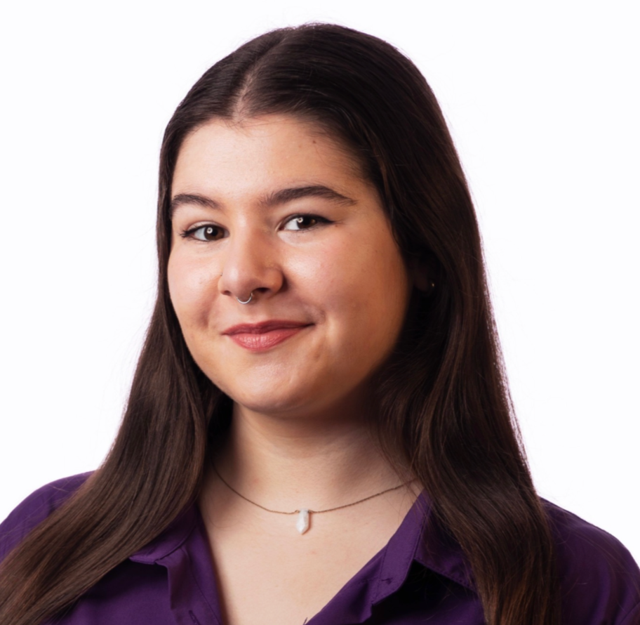 A woman with long brown hair, fair skin, wearing a purple blouse, a small necklace, and a septum piercing, smiling slightly against a white background.