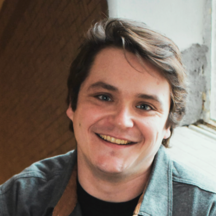 A young man with brown hair smiling at the camera, sitting indoors near a window.