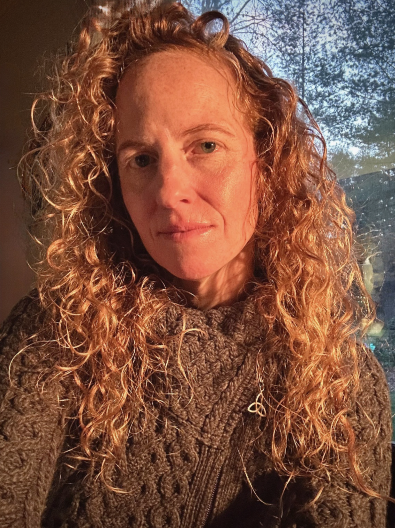 Close-up of a woman with curly red hair, wearing a chunky knit sweater, with a tree and sky in the background.