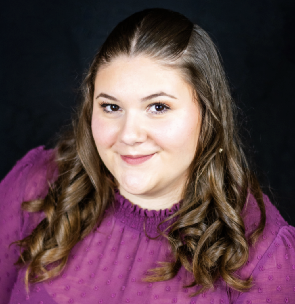Portrait of a young woman with long wavy brown hair wearing a purple textured top, smiling at the camera against a plain black background.
