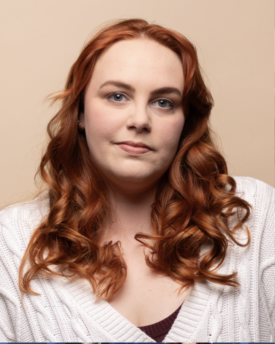 A young woman with long, curly red hair, light skin, wearing a white sweater and small earrings, looking at the camera with a neutral expression.