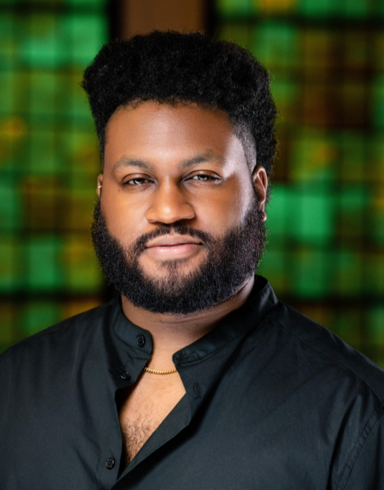 Portrait of a man with a beard and short curly hair, wearing a black shirt and gold chain, standing in front of a green and yellow stained glass background.