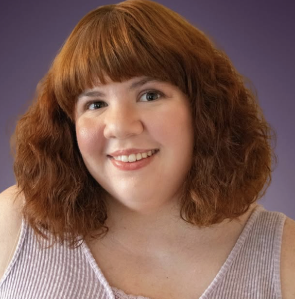 Portrait of a woman with shoulder-length red curly hair, smiling, wearing a striped sleeveless top with a purple background.
