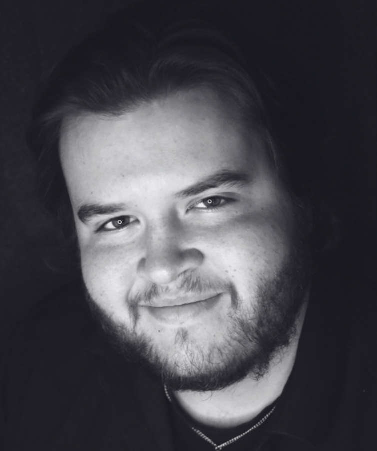 Black and white portrait of a young man with medium-length hair, a beard, and a slight smile looking at the camera.