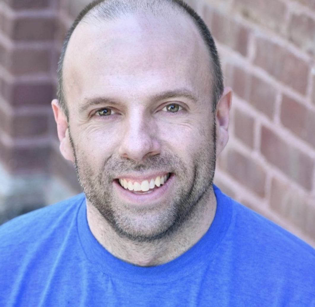 Close-up of a smiling man with short dark hair, wearing a blue shirt, standing outdoors near a brick wall.