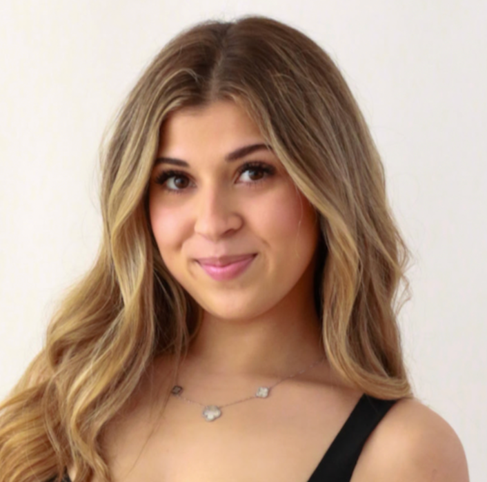Young woman with long wavy blonde hair smiling at the camera, wearing a black top and a necklace with three charms, against a plain white background.