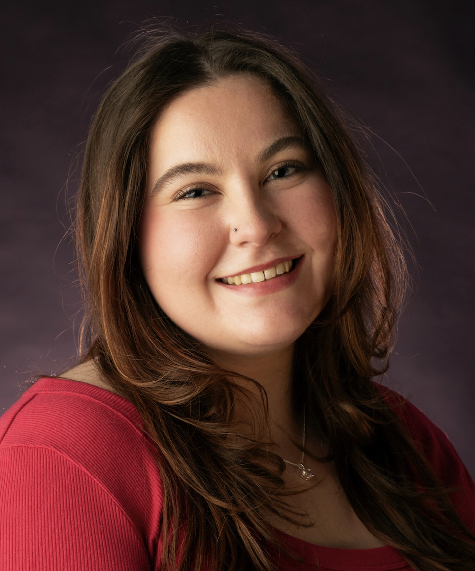 A young woman with long brown hair, smiling, wearing a red top and a necklace, against a dark background.