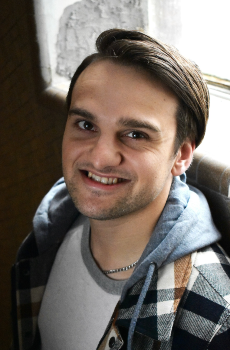A young man with dark hair, smiling and wearing a checkered shirt over a hoodie, sitting near a window.