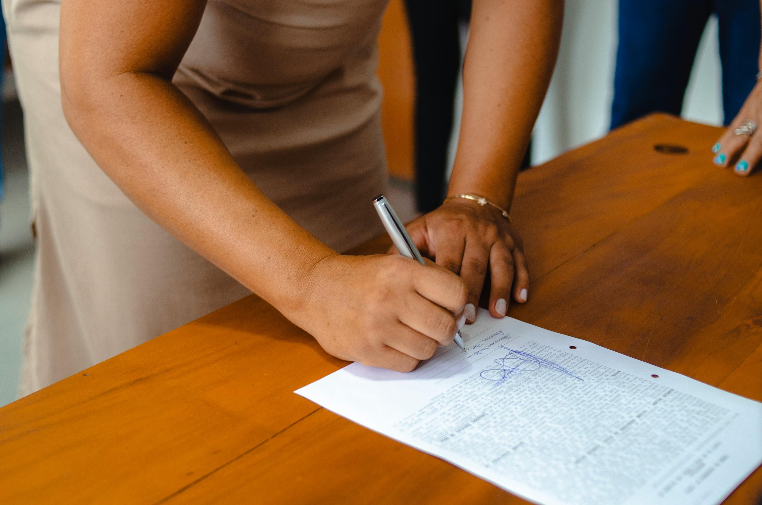 A person signing a document on a wooden table. The person is wearing a beige dress and has turquoise-painted nails.