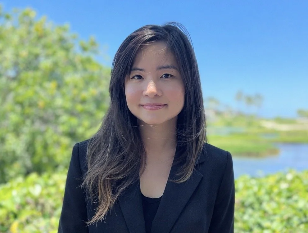 A young woman with long dark hair and a black blazer standing outdoors near a body of water and trees under a clear blue sky.