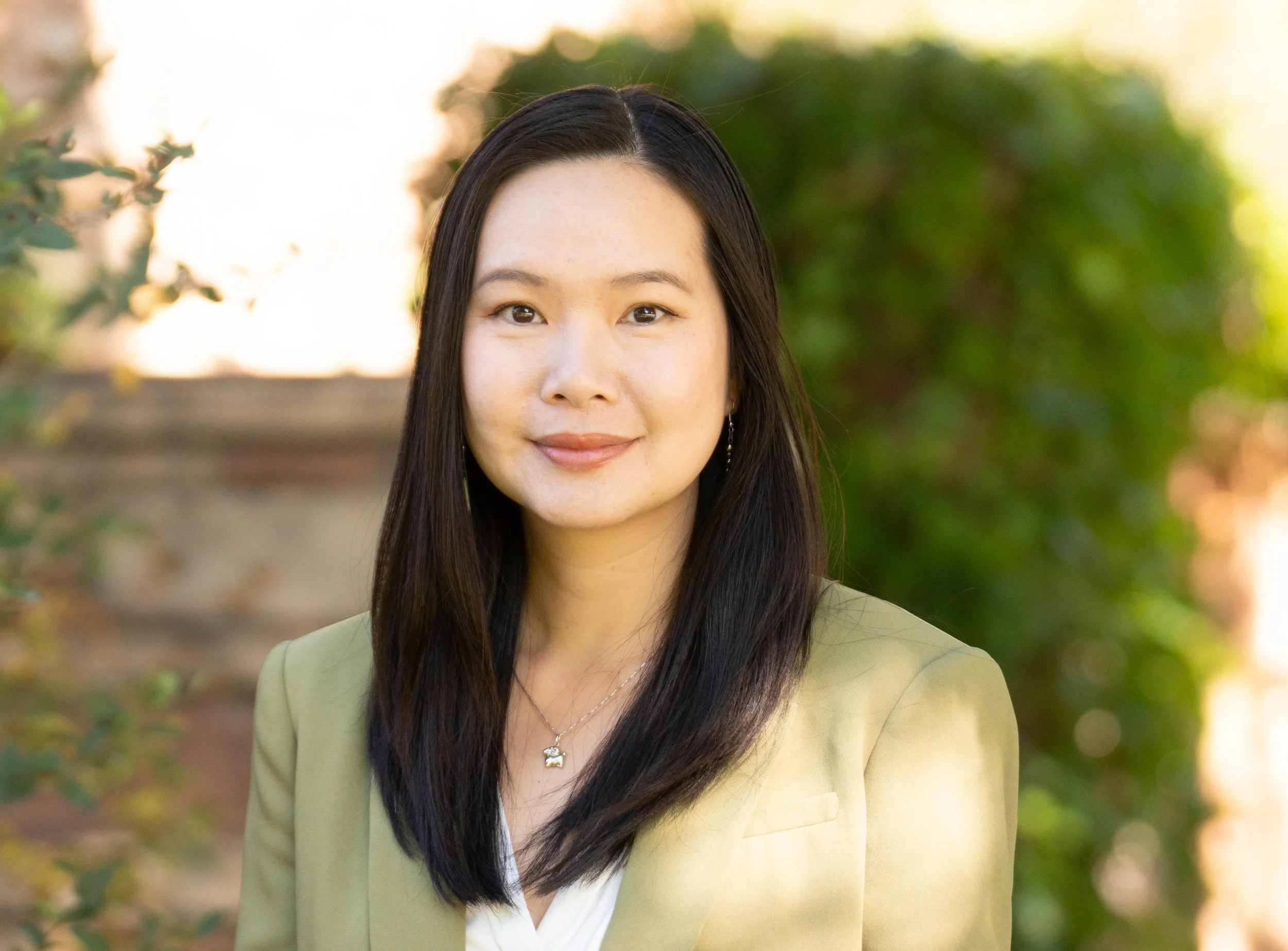 A portrait of a woman with long black hair and a slight smile, standing outdoors with green foliage and sunlight in the background.