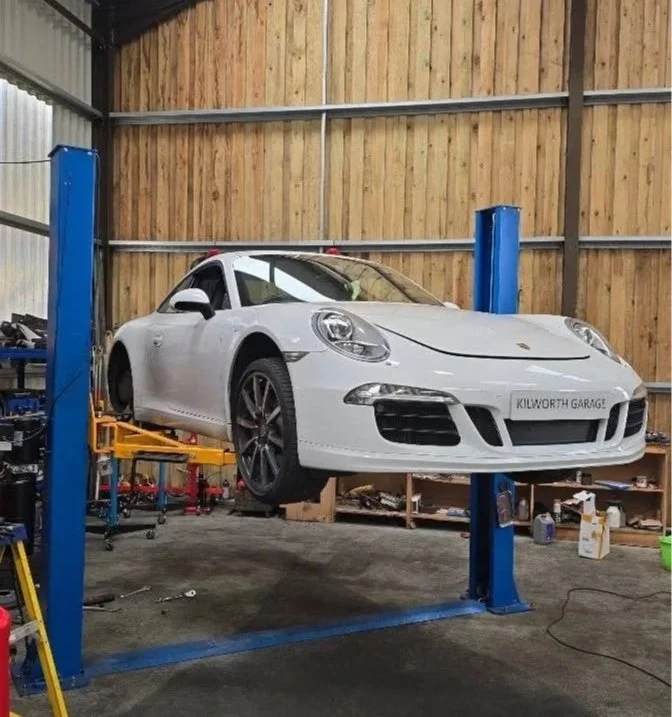 A white car is elevated on a lift inside a garage or workshop with wooden wall paneling. Various tools and supplies are visible on shelves in the background.
