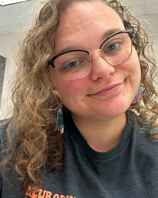 Close-up of a woman with curly hair, glasses, and a black shirt, smiling softly in an indoor setting.