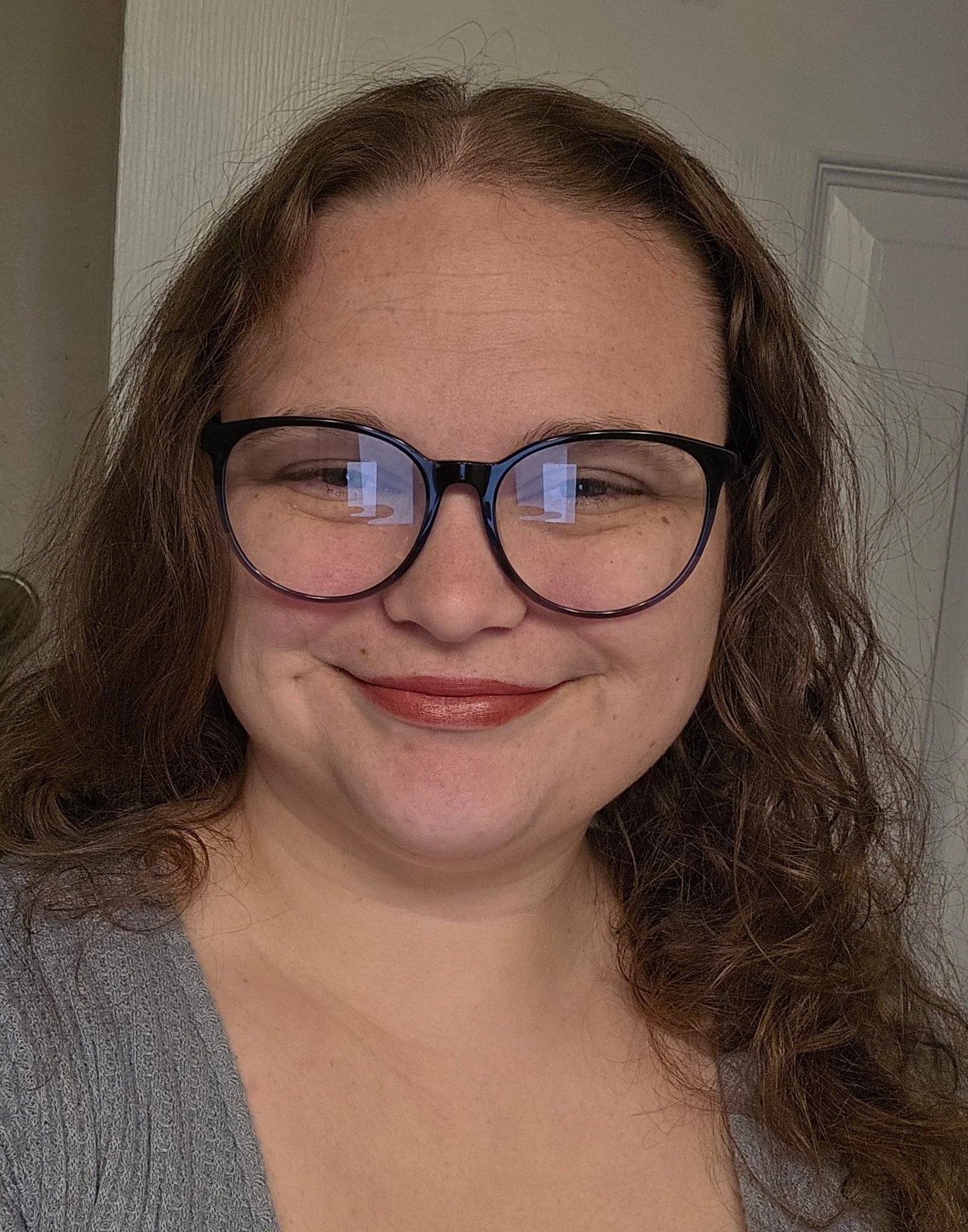 A woman with long curly brown hair wearing glasses, a gray shirt, and red lipstick, smiling at the camera.